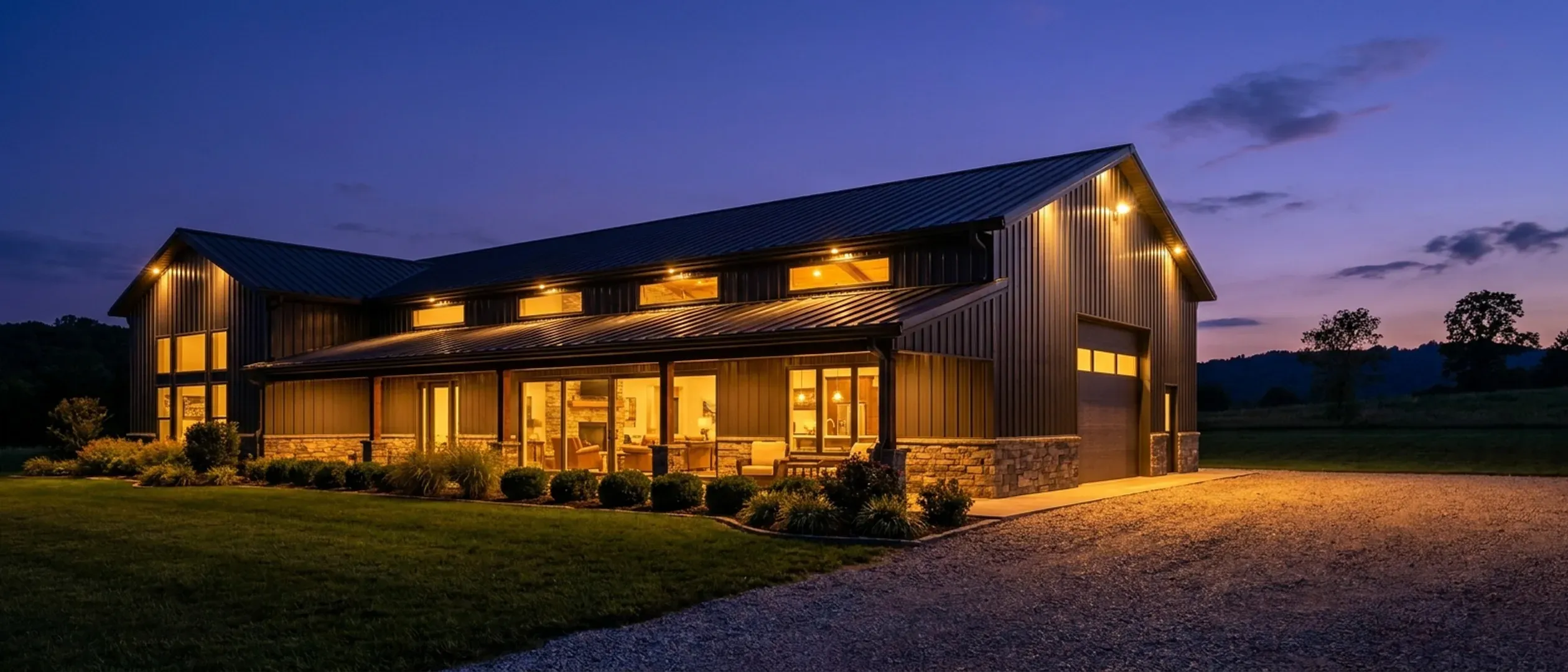 A barndominium at dusk with warm light glowing from every window and exterior lighting along the porch and roofline against a deep blue twilight sky.