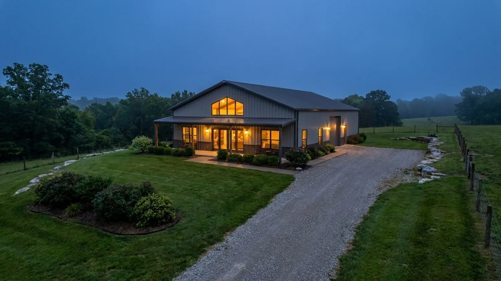 A finished barndominium at dusk with warm interior lighting glowing through its large windows.