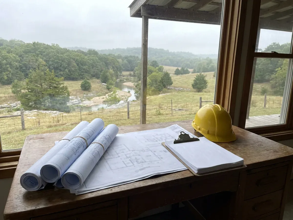 A building permit submittal package on a desk with rolled construction drawings, forms on a clipboard, and a hard hat.