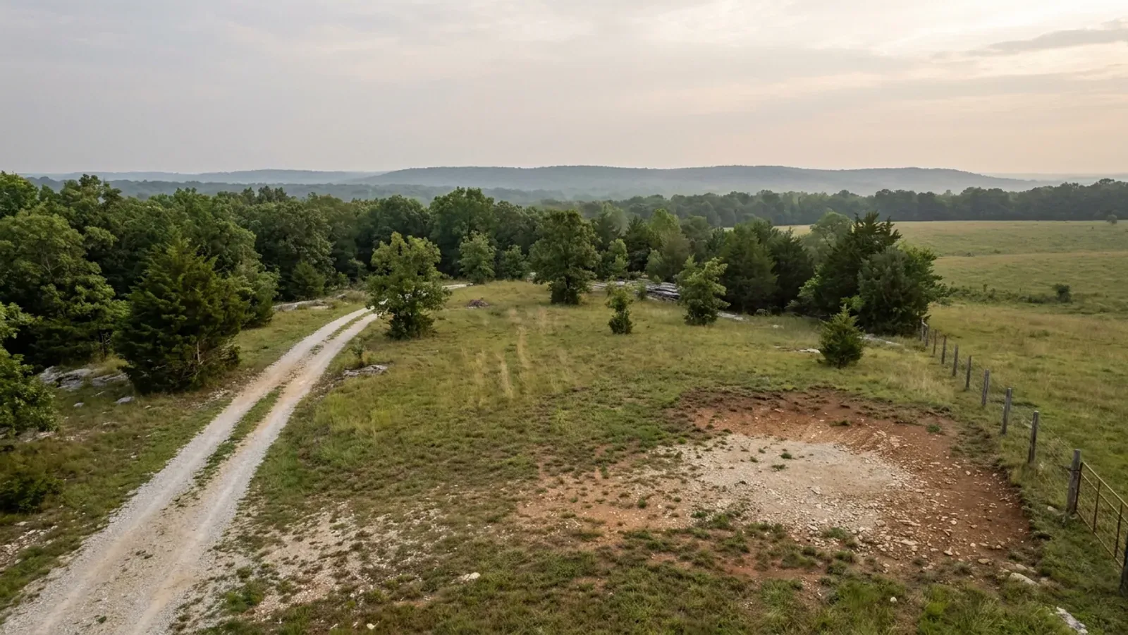 Aerial view of an undeveloped rural lot with cleared grass, a oak and hickory treeline with a few loblolly pines, and a pale gravel access track.